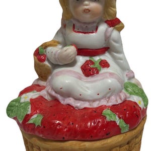 May include: A ceramic figurine of a girl in a pink hat and white dress, holding a basket. The figurine sits atop a red strawberry-shaped base with a woven basket bottom. The figurine is a decorative item.