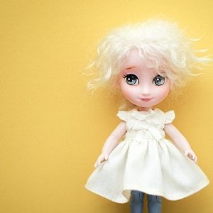 Op de afbeelding: Een witte pop met blond krullend haar, die een witte jurk met een gerimpelde halslijn en een blauwe spijkerbroek draagt. De pop heeft blauwe ogen en staat op een houten vloer.