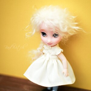 Op de afbeelding: Een witte pop met blond haar en blauwe ogen die een witte jurk draagt. De pop staat op een houten vloer voor een gele muur.