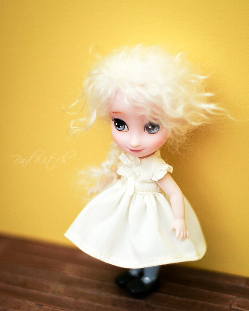 Op de afbeelding: Een witte pop met blond haar en blauwe ogen die een witte jurk draagt. De pop staat op een houten vloer voor een gele muur.