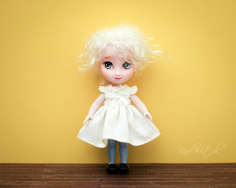 Op de afbeelding: Een witte pop met blond krullend haar, die een witte jurk met een gerimpelde halslijn en een blauwe spijkerbroek draagt. De pop heeft blauwe ogen en staat op een houten vloer.