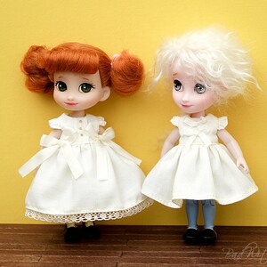 Op de afbeelding: Twee poppen die witte jurken dragen met kantversiering. De pop links heeft rood haar en groene ogen. De pop rechts heeft wit haar en blauwe ogen. Beide poppen staan op een houten vloer.