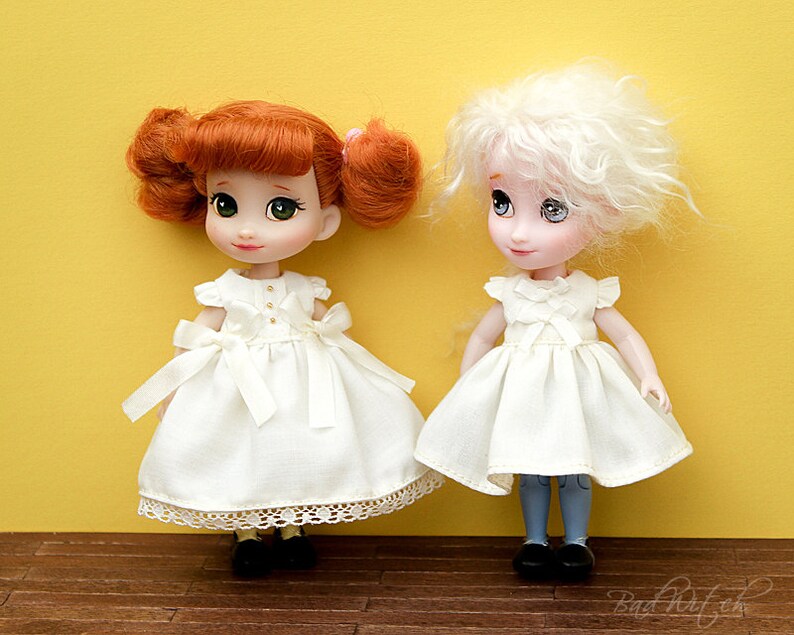 Op de afbeelding: Twee poppen die witte jurken dragen met kantversiering. De pop links heeft rood haar en groene ogen. De pop rechts heeft wit haar en blauwe ogen. Beide poppen staan op een houten vloer.