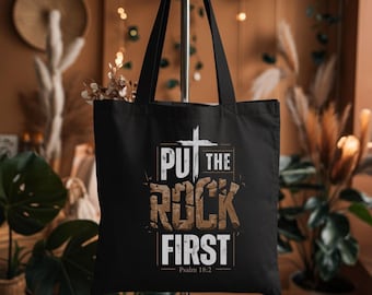 May include: Black tote bag featuring the text "PUT THE ROCK FIRST" in a distressed, textured font, with a cross above the text. The bottom of the bag has the text "Psalm 18:2". The bag has black handles.