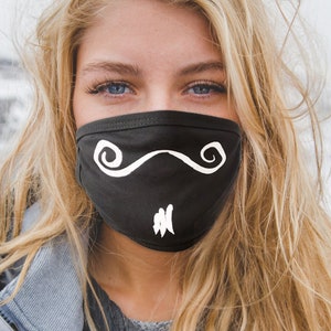 Op de afbeelding: Een vrouw draagt ​​een zwart mondkapje met een wit snorontwerp. Het masker is gemaakt van stof en heeft een comfortabele pasvorm.