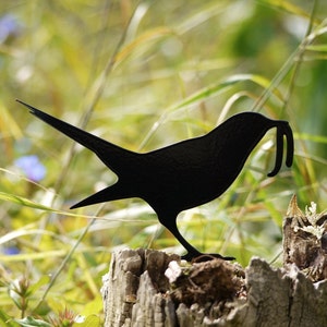 Op de afbeelding: Zwarte metalen silhouet van een vogel met een lange snavel, gepercht op een boomstronk in een grasveld.