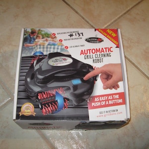 May include: A box for an automatic grill cleaning robot. The black robot has red brushes and blue accents. The box has the text "AUTOMATIC GRILL CLEANING ROBOT" and "AS EASY AS THE PUSH OF A BUTTON!"