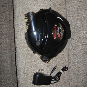 May include: A black, triangular-shaped grill cleaning device with metallic bristles on its sides and a central power button. It includes a black power adapter with a two-prong plug and a thin cord. The device features a warning label with red text.