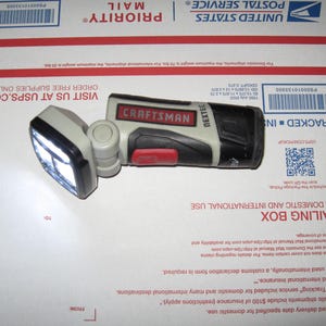 May include: A Craftsman Nextec LED work light, featuring a pivoting head with bright LED lights. The light has a black and gray body with a red button and the Craftsman logo. It is resting on a white surface.