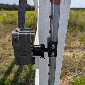 May include: A gray trail camera mounted to a metal fence post with a black bracket. The camera is facing to the right and has a black strap attached to the top.