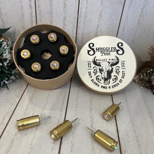 May include: A set of bullet pins in a round cardboard box with the text "Smuggler's Pride Bullet Pins". The pins are gold-colored with iridescent gems. The box lid features a skull graphic. Several loose pins are scattered around the box.