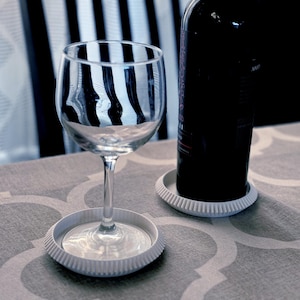May include: A wine glass and a bottle of red wine sit on a patterned tablecloth. The wine bottle rests on a white coaster, and the wine glass sits on a matching coaster. The background includes a black chair and a window.