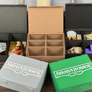 May include: A collection of rectangular storage boxes for Arkham Horror: The Card Game. The set includes open boxes in black and tan, and closed boxes in gray and green, each featuring the white printed text Arkham Horror The Card Game.