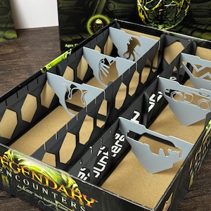 May include: A black cardboard game box insert featuring multiple slots separated by light gray dividers. Each divider has a unique die-cut silhouette shape. The box exterior is printed with the text Legendary Encounters An Alien Deck Building Game.