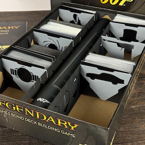 May include: A Legendary James Bond deck building game box with internal dividers. The black box features the 007 logo and contains multiple card slots with grey dividers displaying silhouettes of iconic imagery like a car and a gun barrel.