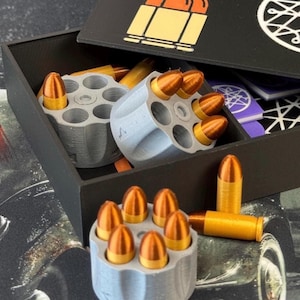 May include: A black box with a lid featuring a gold bullet graphic. Inside, gray holders contain copper-colored bullets. Additional bullets are scattered around the holders. The box sits on a patterned surface.