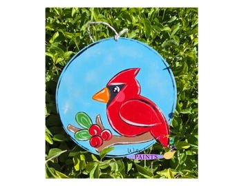 May include: A round wooden sign with a blue background and a painted cardinal perched on a branch with red berries. The sign has the text "Wendy's Paints" at the bottom.