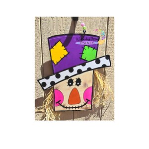May include: A wooden scarecrow face with a purple hat with yellow and green patches, a white and black polka dot brim, and a straw beard. The scarecrow has orange and pink painted features and a stitched smile.