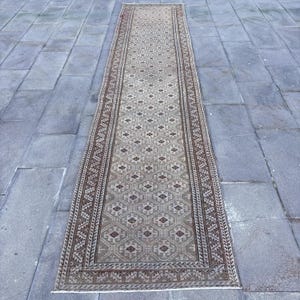 Peut inclure: Un long tapis de couloir étroit présentant un motif géométrique répétitif en losanges dans des tons beige, marron et gris foncé. Le champ central est encadré par une bordure décorative aux motifs linéaires complexes et symétriques.