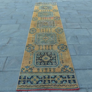 May include: A long, narrow runner rug featuring a mustard yellow background with a series of repeating square medallions in shades of teal, navy blue, and orange. The design includes intricate geometric borders and floral motifs throughout the piece.