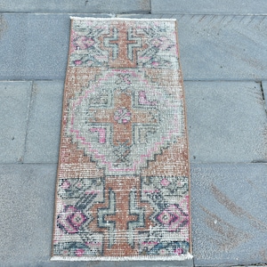 May include: A rectangular woven rug featuring a geometric pattern in shades of terracotta, muted gray, and vibrant pink. The design includes a central cross motif surrounded by intricate borders and small fringe details at the top and bottom edges.