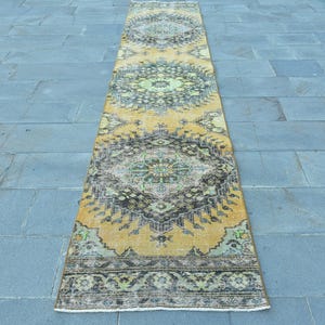Peut inclure: Un tapis de couloir rectangulaire de style vintage avec un aspect vieilli. Le tapis présente un motif de médaillon central dans des tons de jaune, noir et vert. Il est posé sur une surface en pierre grise.