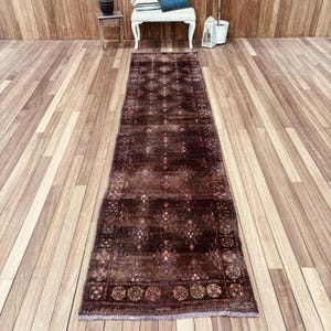 Puede incluir: Una alfombra pasillera larga y rectangular con un color base marrón oscuro y un patrón repetitivo de pequeños motivos en forma de diamante en tonos apagados de rojo, naranja y crema. La alfombra tiene un borde con flecos y textura oscura.