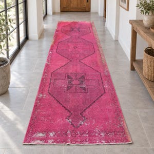 Puede incluir: Una alfombra de pasillo larga y rectangular en un tono rosa intenso y vibrante. La alfombra presenta una serie de patrones geométricos oscuros en forma de diamante a lo largo del centro, con áreas desgastadas y descoloridas.