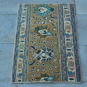 May include: A rectangular rug featuring a tan central field decorated with stylized floral motifs in shades of teal, blue, and cream. The rug is framed by multiple decorative borders in blue, tan, and cream, showcasing intricate geometric patterns.