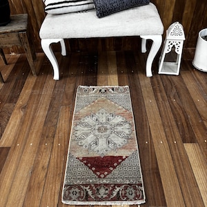 May include: A rectangular area rug featuring a central geometric medallion in shades of beige, gray, and deep red. The rug displays intricate border patterns and a repeating grid motif, with a weathered texture and short fringe along the edges.