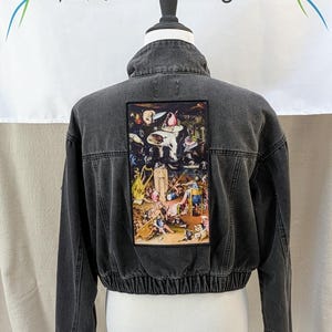 May include: A charcoal gray cropped denim jacket featuring a large rectangular patch on the back with a detailed reproduction of Hieronymus Bosch's The Garden of Earthly Delights. The jacket has a high collar, long sleeves, and an elasticized hem.