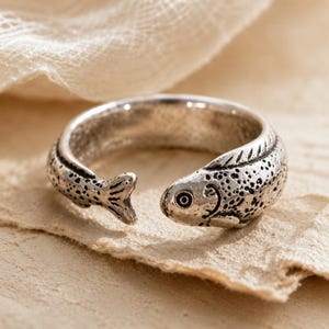 May include: An open-style silver-toned ring shaped like a fish. The metal surface features a textured, speckled pattern across the body, with a detailed head showing an eye and a tail fin at the opposite end of the band, creating a continuous wrap design.