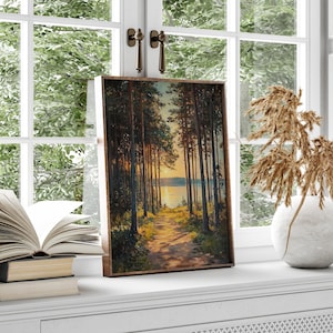 May include: A framed landscape painting shows a path through a forest towards a lake at sunset. The artwork features tall trees with a warm, golden light. The frame is a dark brown colour. The painting is displayed on a windowsill.