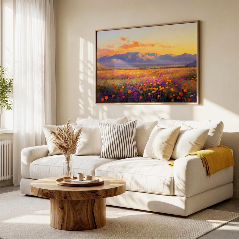 May include: A living room scene featuring a large landscape painting with a sunset over a field of flowers and mountains. A cream-colored sofa with pillows and a wooden coffee table complete the decor.