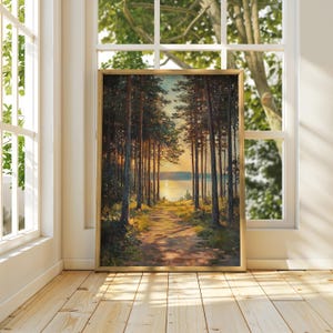May include: A framed landscape painting shows a path through a forest leading to a lake at sunset. Tall trees line the path, with sunlight filtering through the branches. The painting is in a gold frame, displayed near a window, with natural light.