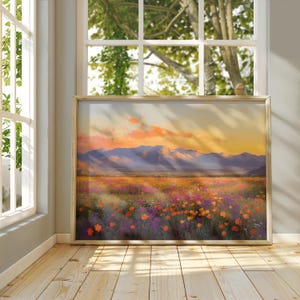 May include: A framed landscape painting depicts a field of wildflowers under a sunset sky. The artwork features orange, purple, and yellow flowers, with mountains in the background. The frame is a light gold color.