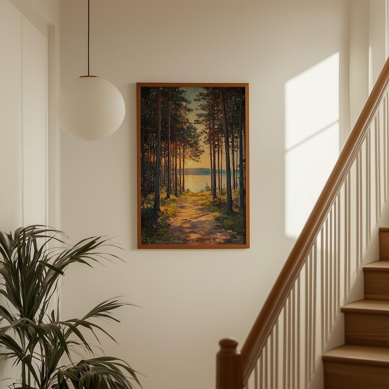 May include: A framed landscape painting shows a path through a forest leading to a lake. The artwork features tall trees, a dirt path, and a warm, golden light illuminating the scene. The frame is a warm brown colour.