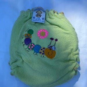 May include: A light green cloth diaper with a colorful embroidered caterpillar and pink flower design. The diaper has a tag that reads "Large 18-30 lbs".