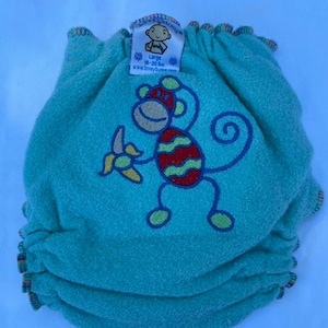 May include: A teal cloth diaper with a monkey graphic. The monkey has a red and yellow striped torso, a tan face, and is holding a yellow banana. The diaper has a tag that says "Large 18-30 lbs."