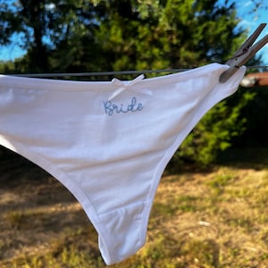 Puede incluir: Tanga blanco con un pequeño lazo y la palabra "Bride" bordada en azul claro. La ropa interior está colgada en un tendedero con una pinza de madera, sobre un fondo borroso de follaje verde y cielo azul.