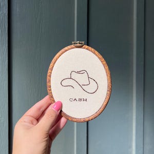 May include: An oval embroidery hoop with a faux wood grain frame containing off-white fabric. The fabric features a dark brown stitched outline of a cowboy hat and the word CASH embroidered in dark brown thread below the hat design.