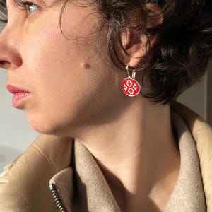 Puede incluir: Un par de pendientes de plata con abalorios redondos bordados en rojo y blanco. Los abalorios presentan un patrón geométrico.