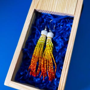 Puede incluir: Pendientes de cuentas en una caja de madera. Los pendientes presentan un degradado de amarillo, naranja y rojo, con la parte superior de cuentas blancas y ganchos plateados. Se encuentran sobre papel de seda azul.