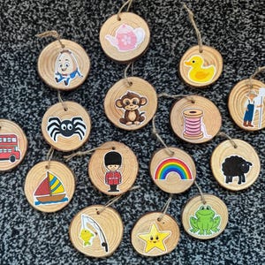 May include: A selection of wooden disc ornaments, each featuring a different colourful illustration. Designs include a London bus, a spider, a sailboat, a rainbow, a frog, and a star. Each has a jute string.