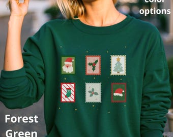 Könnte beinhalten: Forstgrünes Sweatshirt mit sechs weihnachtlichen Briefmarkenmotiven. Die Briefmarken zeigen den Weihnachtsmann, Stechpalme, einen Weihnachtsbaum, einen Strumpf, einen Stechpalmenzweig und einen Weihnachtsmannhut. Der Text lautet "More color options" und "Forest Green Color".