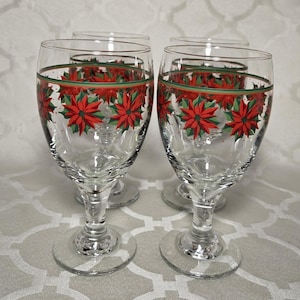 Op de afbeelding: Vier heldere wijnglazen met een feestelijk poinsettia-ontwerp. Elk glas heeft een rood poinsettia-patroon met groene bladeren en een groene rand. De glazen hebben een klassieke steel en voet.