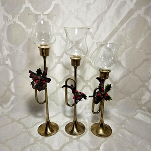 May include: Three brass trumpet-shaped candle holders of varying heights, each topped with a clear glass hurricane shade. The brass stems are decorated with small festive accents featuring green and red plaid ribbons, pinecones, and red berries.