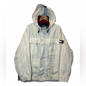 May include: A light beige hooded jacket with multiple front pockets and a navy blue and red interior lining. The jacket features a Tommy Hilfiger logo patch on the sleeve. The jacket has a zipper closure and a drawstring hood.