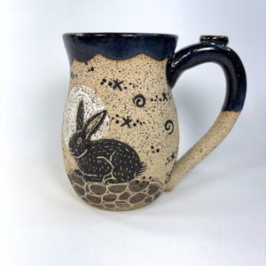 May include: A ceramic mug with a speckled beige body and a dark blue glazed rim and handle. The mug features a black etched illustration of a rabbit sitting on a stone-like base against a white circular moon, surrounded by small star and swirl motifs.