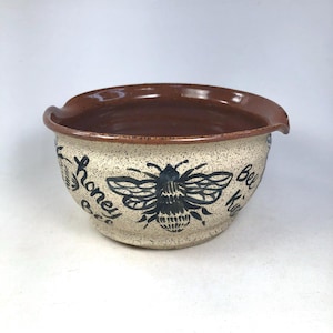 May include: A ceramic bowl with a speckled beige exterior and a dark brown interior. The bowl features a large black bee illustration and the words "honey bee kind" in a handwritten font. The rim has a slight spout.